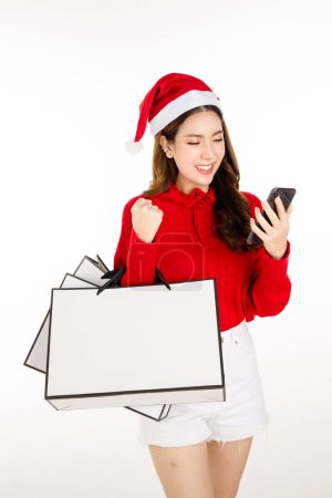 Foto de Alegre mujer asiática atractiva con un vestido rojo con un sombrero de Santa es la celebración de un bolsas de compras sobre fondo blanco aislado. Hermosa dama con el pelo largo y negro está utilizando el teléfono celular de compras en línea. - Imagen libre de derechos