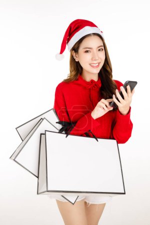 Foto de Alegre mujer asiática atractiva con un vestido rojo con un sombrero de Santa es la celebración de un bolsas de compras sobre fondo blanco aislado. Hermosa dama con el pelo largo y negro está utilizando el teléfono celular de compras en línea. - Imagen libre de derechos