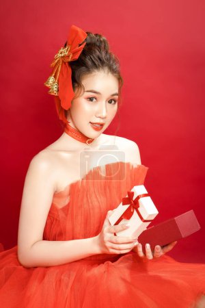 Junge asiatische hübsche Frau Modell in einem noblen stilvollen roten Luxuskleid auf rotem Hintergrund isoliert. Aufgeregt hält eine Frau eine Schachtel in der Hand. Frohes neues Jahr.
