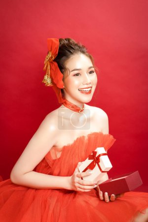 Junge asiatische hübsche Frau Modell in einem noblen stilvollen roten Luxuskleid auf rotem Hintergrund isoliert. Aufgeregt hält eine Frau eine Schachtel in der Hand. Frohes neues Jahr.