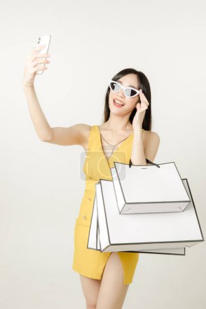 Foto de Retrato de mujer asiática confiada en un vestido amarillo sensual con gafas de sol y llevar una bolsa de paquetes es divertirse compras después de comprar con teléfono móvil aislado sobre fondo blanco. - Imagen libre de derechos