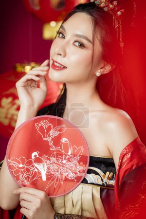 Feliz año nuevo chino. Hermosa mujer asiática usando cheongsam qipao vestido tradicional mira a la cámara con gesto de felicitación sosteniendo ventilador aislado sobre fondo rojo.