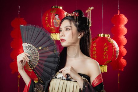 Frohes chinesisches neues Jahr. Schöne Asiatin in traditionellem Cheongsam-Qipao-Kleid blickt in die Kamera mit Geste der Gratulation hält Fan isoliert auf rotem Hintergrund.