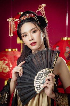Foto de Feliz año nuevo chino. Hermosa mujer asiática usando cheongsam qipao vestido tradicional mira a la cámara con gesto de felicitación sosteniendo ventilador aislado sobre fondo rojo. - Imagen libre de derechos