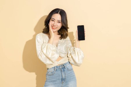 Photo for Asian happy woman with perfect body show blank screen of mobile phone in studio. Luxury fashionable female model in sexy dress with on beige isolated background. E-commerce, Online shopping Concept. - Royalty Free Image