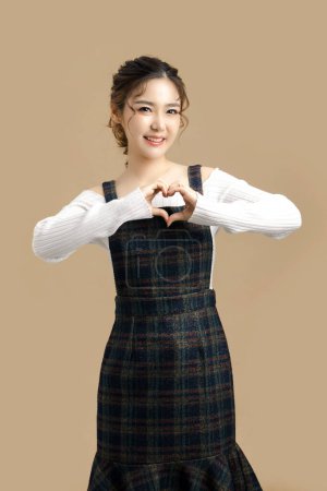 Photo for Happy beautiful Asian model showing heart sign with copy space for separate products on beige background. - Royalty Free Image