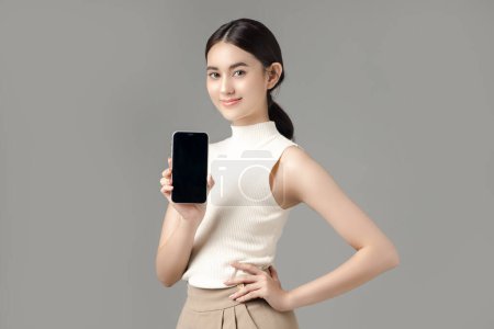 Photo for Confident Asian woman holding phone show blank screen and looking at camera isolated on gray background. Portrait of a beautiful girl in the studio. - Royalty Free Image