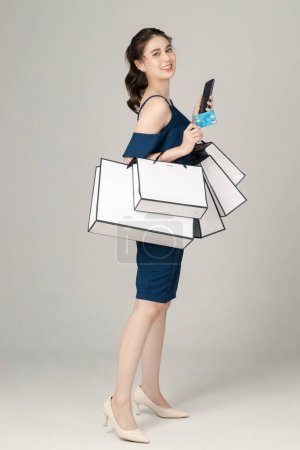 Young energetic Asian woman holding credit crad, mobile phone with shopping bags on gray background. Portrait of pretty girl in studio. cashless society Concept.