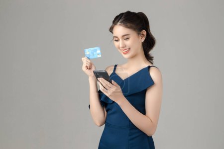 Young energetic Asian woman holding credit card with blank screen mobile phone on gray background. Portrait of pretty girl in studio. cashless society Concept.