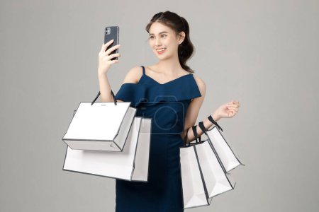 Jeune femme asiatique énergique tenant téléphone portable avec des sacs à provisions et selfie sur fond gris. Portrait de jolie fille en studio. société sans numéraire Concept.