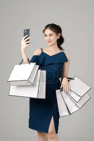 Photo for Young energetic Asian woman holding mobile phone with shopping bags and selfie on gray background. Portrait of pretty girl in studio. cashless society Concept. - Royalty Free Image