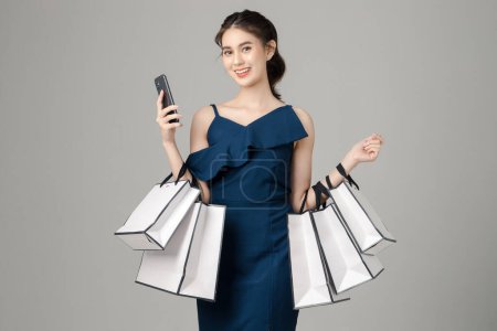 Young energetic Asian woman holding blank screen mobile phone with shopping bags on gray background. Portrait of pretty girl in studio. cashless society Concept.