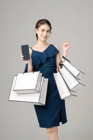 Young energetic Asian woman holding blank screen mobile phone with shopping bags on gray background. Portrait of pretty girl in studio. cashless society Concept.