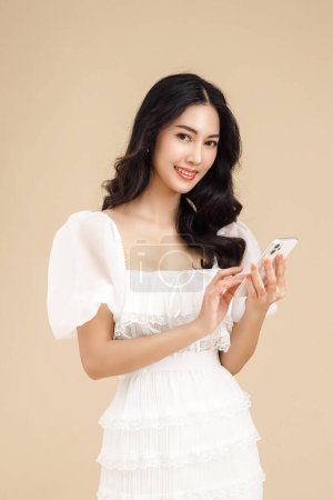 Téléchargez les photos : Jeune femme asiatique à la mode utilisant smartphone debout sur fond beige isolé se sentant heureux. Achats paiement en ligne avec téléphone mobile. Femme montrant un téléphone cellulaire à écran vierge. - en image libre de droit