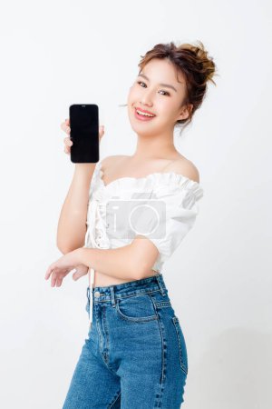 Téléchargez les photos : Belle femme asiatique à la mode montrant écran vierge téléphone mobile sur fond blanc isolé et l'espace de copie. Achats paiement en ligne. sourire de bonne humeur. - en image libre de droit