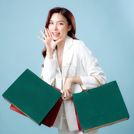 Photo for Half body of beautiful Asian Business woman wearing white suit holding shopping bag and mobile phone on isolated blue background. Blank screen smartphone concept. - Royalty Free Image