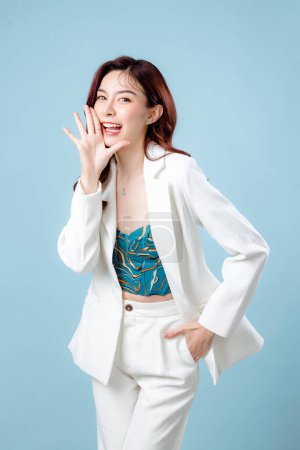 Photo for Half body of confident elegant beautiful Asian Business woman wearing white suit open mouths raising hands screaming announcement on isolated blue background. Young shopaholic female on copy space. - Royalty Free Image