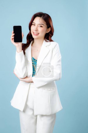 Téléchargez les photos : Demi-corps de confiant élégant belle femme d'affaires asiatique portant costume blanc sourire et tenant téléphone mobile sur fond bleu isolé. Concept de smartphone à écran blanc. - en image libre de droit