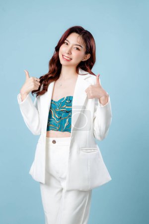 Photo for Half body of confident elegant beautiful Asian Business woman wearing white suit smiling and showing thumbs up on isolated blue background. Young shopaholic female on copy space. - Royalty Free Image