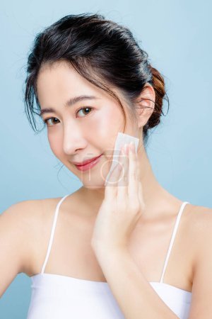 Photo for Beautiful Asian woman with perfect clear skin. Happy girl model with fresh hydrated facial wipe her face on isolated blue background. Cosmetology , beauty and spa, wellness, Plastic surgery. - Royalty Free Image