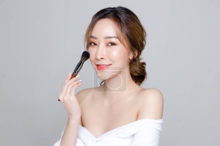 Photo for Asian woman with a beautiful face and Perfect clean fresh skin. Portrait of female model using brush for makeup on Grey isolated background. Cosmetology, Body care, plastic surgery, beauty and spa. - Royalty Free Image