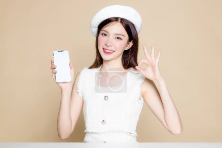 Photo for Cute Asian woman with perfect skin and showing blank display. Pretty girl model wearing white beret and natural makeup on beige background. Cosmetology, beauty and spa, wellness, Plastic surgery. - Royalty Free Image