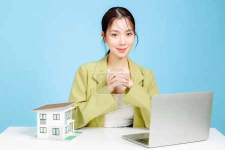Photo for Young Asian business woman wearing casual suit with a small model house with laptop on blue background. Portrait of confident female investment agency using social media contacting with buyer. - Royalty Free Image