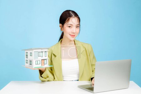 Photo for Young Asian business woman wearing casual suit holding a small model house with laptop on blue background. Portrait of confident female investment agency using social media contacting with buyer. - Royalty Free Image