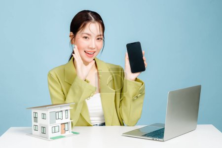 Photo for Young Asian business woman wearing casual suit with a small model house with laptop and holding mobile phone on blue background. Portrait of confident female investment agency using social media. - Royalty Free Image