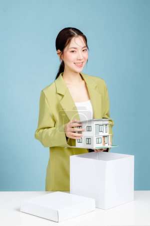Foto de Joven mujer de negocios de éxito inversionista en traje casual sosteniendo modelo de casa en escritorio de oficina blanco con PC portátil aislado en retrato de estudio de fondo azul pastel. Concepto de logro - Imagen libre de derechos