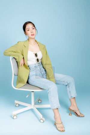 Photo for Young beautiful attractive Asian woman in casual green fashion jacket and sunglasses sitting on armchair isolated on pastel blue background. Female Consumerism, lifestyle concept. - Royalty Free Image