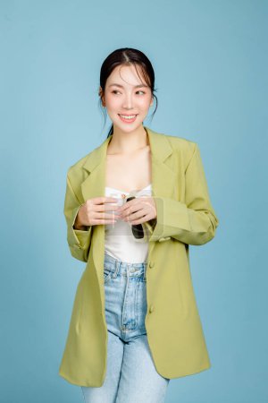 Photo for Young beautiful attractive Asian woman in casual green fashion jacket and sunglasses isolated on pastel blue background. Female Consumerism, lifestyle concept. - Royalty Free Image