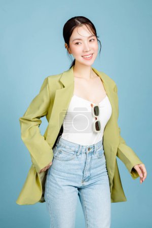 Foto de Joven hermosa mujer asiática atractiva en chaqueta casual de moda verde y gafas de sol aisladas sobre fondo azul pastel. Consumismo femenino, concepto de estilo de vida. - Imagen libre de derechos