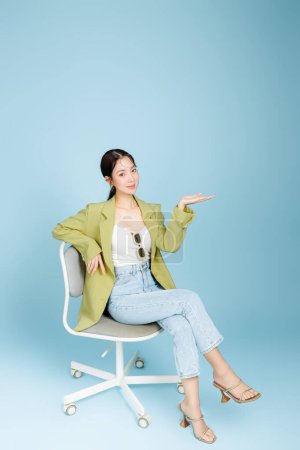 Photo for Young beautiful attractive Asian woman in casual green fashion jacket and sunglasses sitting on armchair showing copy space isolated on pastel blue background. Female Consumerism, lifestyle concept. - Royalty Free Image