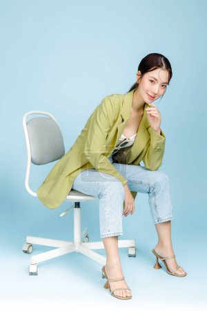 Foto de Joven hermosa mujer asiática atractiva en chaqueta casual de moda verde y gafas de sol sentados en sillón aislado sobre fondo azul pastel. Consumismo femenino, concepto de estilo de vida. - Imagen libre de derechos