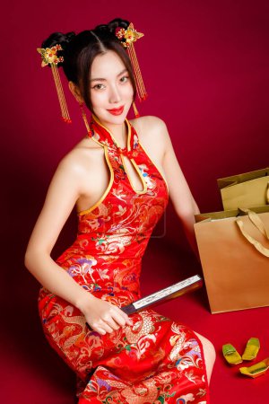 Photo for Beautiful Asian woman with clean fresh skin wearing cheongsam dress holding fan sitting with gold shopping bags on red background. Portrait of female model in studio. Happy Chinese new year. - Royalty Free Image