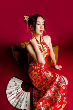 Photo for Beautiful Asian woman with clean fresh skin wearing cheongsam dress holding fan sitting with gold shopping bags on red background. Chinese text means looking for spring and happy to see plum. - Royalty Free Image
