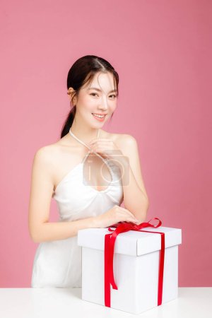 Photo for Young Asian woman gathered in ponytail with natural makeup on face and clean fresh skin wearing white camisole on isolated pink background. Portrait of cute female model with gift box in studio. - Royalty Free Image