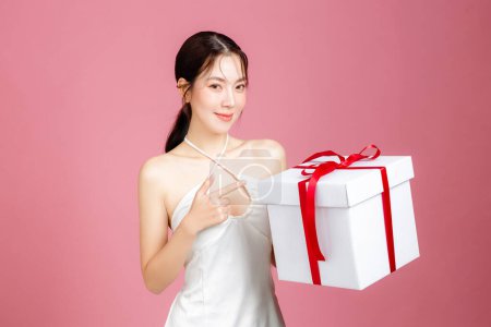 Photo for Young Asian woman gathered in ponytail with natural makeup on face and clean fresh skin wearing white camisole on isolated pink background. Portrait of cute female model with gift box in studio. - Royalty Free Image