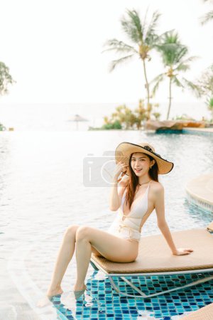 Photo for Portrait of gorgeous Asian woman model beauty skincare suntan posing sunbathing tanning in white one piece swimsuit and straw hat relaxing at infinity pool luxury resort. Spa, wellness, laser. - Royalty Free Image