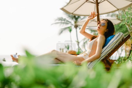 Téléchargez les photos : Portrait de magnifique femme asiatique modèle beauté soin bronzage couché sur une chaise de plage en maillot de bain une pièce blanc relaxant à la station balnéaire d'été. Spa, bien-être, laser. - en image libre de droit