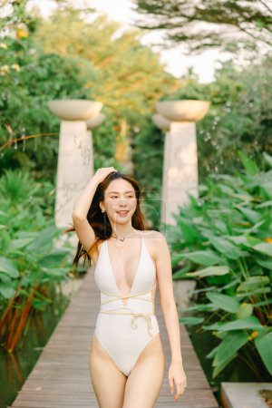 Téléchargez les photos : Portrait de magnifique asiatique femme modèle beauté soin bronzage marche dans le jardin tropical en blanc maillot de bain une pièce relaxant à la station balnéaire d'été. Spa, bien-être, laser. - en image libre de droit