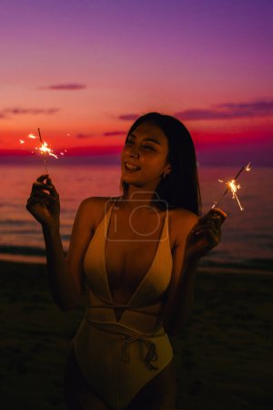 Photo for Side view of happy perfect slim body beautiful Asian woman in swimsuit silhouette relaxing with firework at sunset enjoying perfect beach holiday vacation. Summer travel. - Royalty Free Image