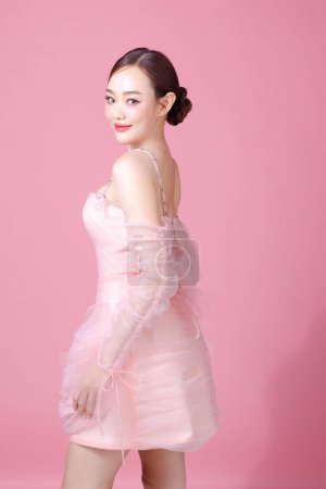 Téléchargez les photos : Fascinant plaisir joyeuse jeune femme de l'Asie porte douce robe de fleur rose pose geste isolé sur fond rose clair pastel plaine beau portrait de dame en studio. - en image libre de droit