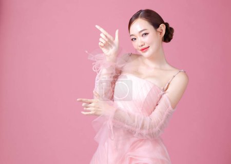 Téléchargez les photos : Fascinant plaisir joyeuse jeune femme de l'Asie porte robe de fleur rose doux pose geste pointant du doigt pour copier l'espace isolé sur fond rose clair pastel ordinaire Portrait de dame en studio. - en image libre de droit