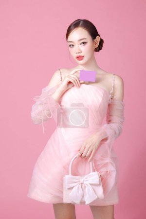 Photo for Fascinating fun joyful young woman of Asian wears sweet pink flower dress hold in hand credit bank card cover isolated on plain pastel light pink background Beautiful lady portrait in studio. - Royalty Free Image