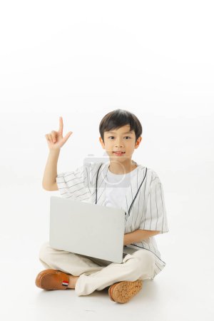 Téléchargez les photos : Heureux petit garçon asiatique assis en tapant ordinateur portable tout en pensant isolé sur fond blanc, concept d'apprentissage en ligne, En regardant la caméra et la composition complète du corps. - en image libre de droit