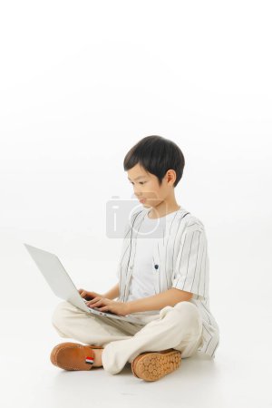 Foto de Feliz asiático pequeño niño sentado escribiendo portátil aislado sobre fondo blanco, concepto de aprendizaje en línea, mirando a la cámara y la composición de cuerpo completo. - Imagen libre de derechos