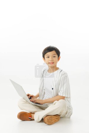 Téléchargez les photos : Heureux petit garçon asiatique assis en tapant ordinateur portable isolé sur fond blanc, concept d'apprentissage en ligne, En regardant la caméra et la composition complète du corps. - en image libre de droit