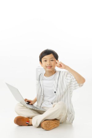 Téléchargez les photos : Heureux petit garçon asiatique assis en tapant ordinateur portable tout en pensant isolé sur fond blanc, concept d'apprentissage en ligne, En regardant la caméra et la composition complète du corps. - en image libre de droit